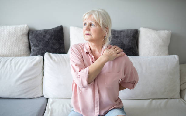 An elderly woman sits on a couch, holding her shoulder and looking pensive.